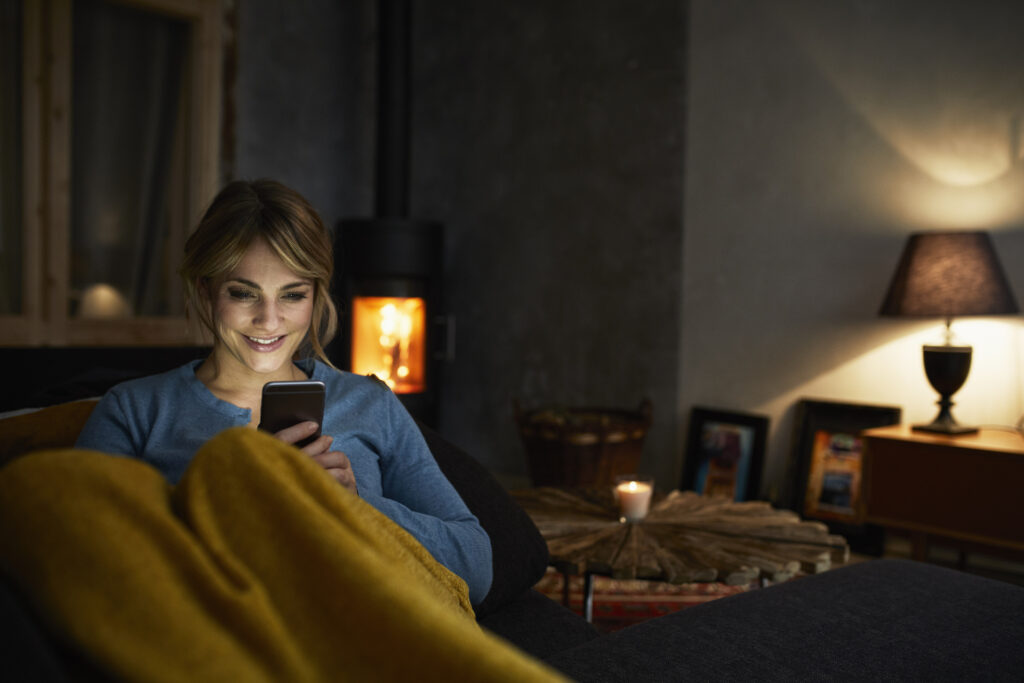 Portrait of smiling woman with smartphone relaxing on couch in the evening - Tinn Energi & Fiber - Tinn Energi & Fiber Eva smarthus produkter